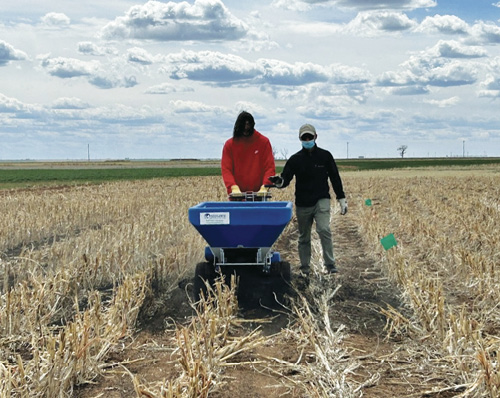 Two people in a plot field operate a blue biochar spreader under a cloudy sky.