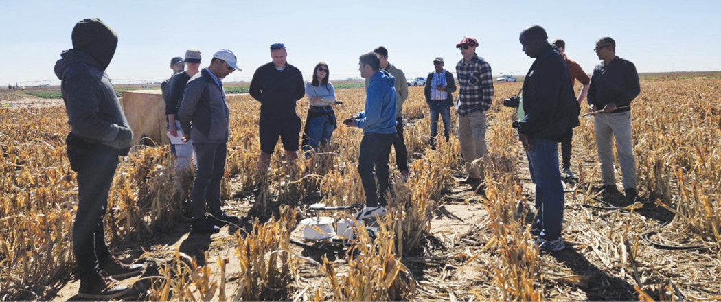 Group of people standing in a harvested crop field, listening to a person speaking during an outdoor field demonstration.