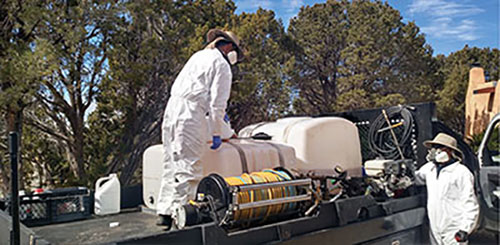 Photograph of two people wearing respirators, Tyvek coveralls, and gloves with herbicide tanks on a flatbed truck.