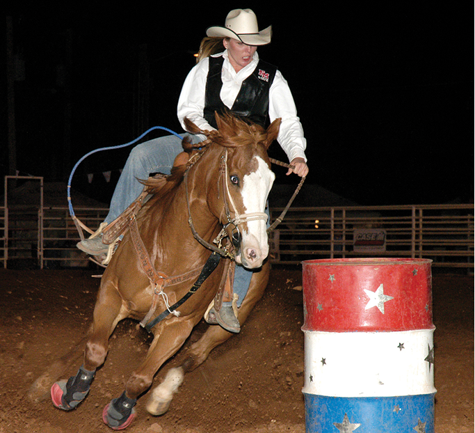 A cowgirl competes in barrel racing, skillfully guiding her horse around a red, white, and blue barrel under nighttime lights.