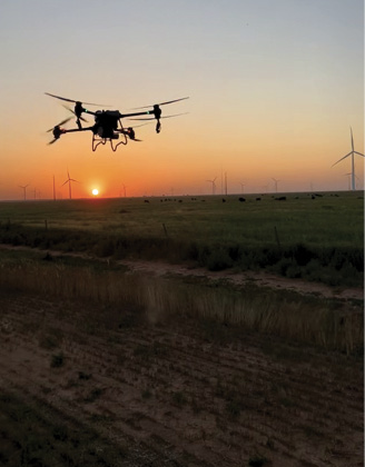 Drone flying over a field at sunset with wind turbines in the background.