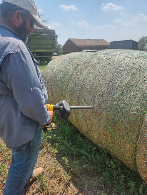 A person is holding a drill attached to a sampling probe, positioned to take samples from large, round hay bales. The drill is shown in action, with the probe extending into a bale. The focus of the image is on the proper sampling technique, highlighting the correct location on the bale for effective sampling.