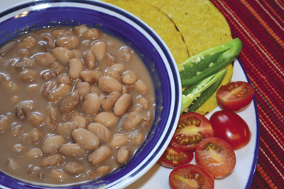 Photograph of a bowl of pinto beans with corn tortillas, jalapeños, and tomatoes.