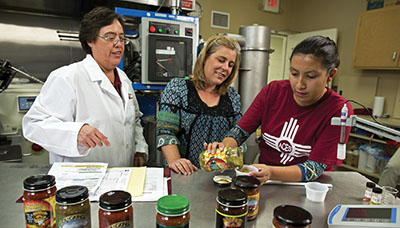 Photograph of three people in a commercial kitchen. One person is pouring salsa from a jar into a small dish.