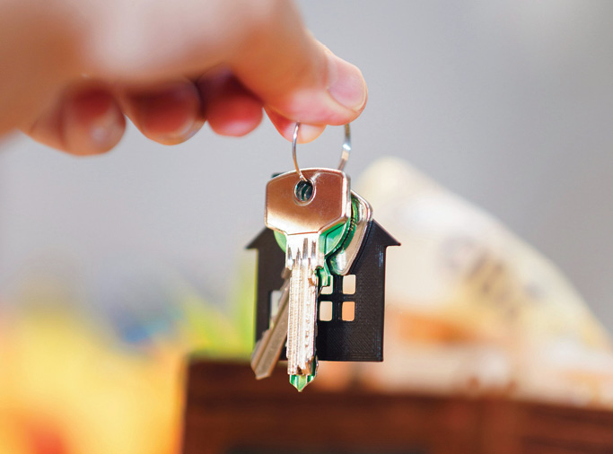 Close-up of a hand holding a keyring with house keys and a small house-shaped keychain, with a softly blurred home interior in the background.