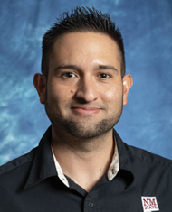 Studio headshot of a smiling man with short dark hair wearing a black collared shirt against a blue mottled backdrop.