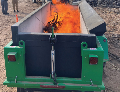 Rear view of a green industrial burn box with flames burning inside at an outdoor work site.
