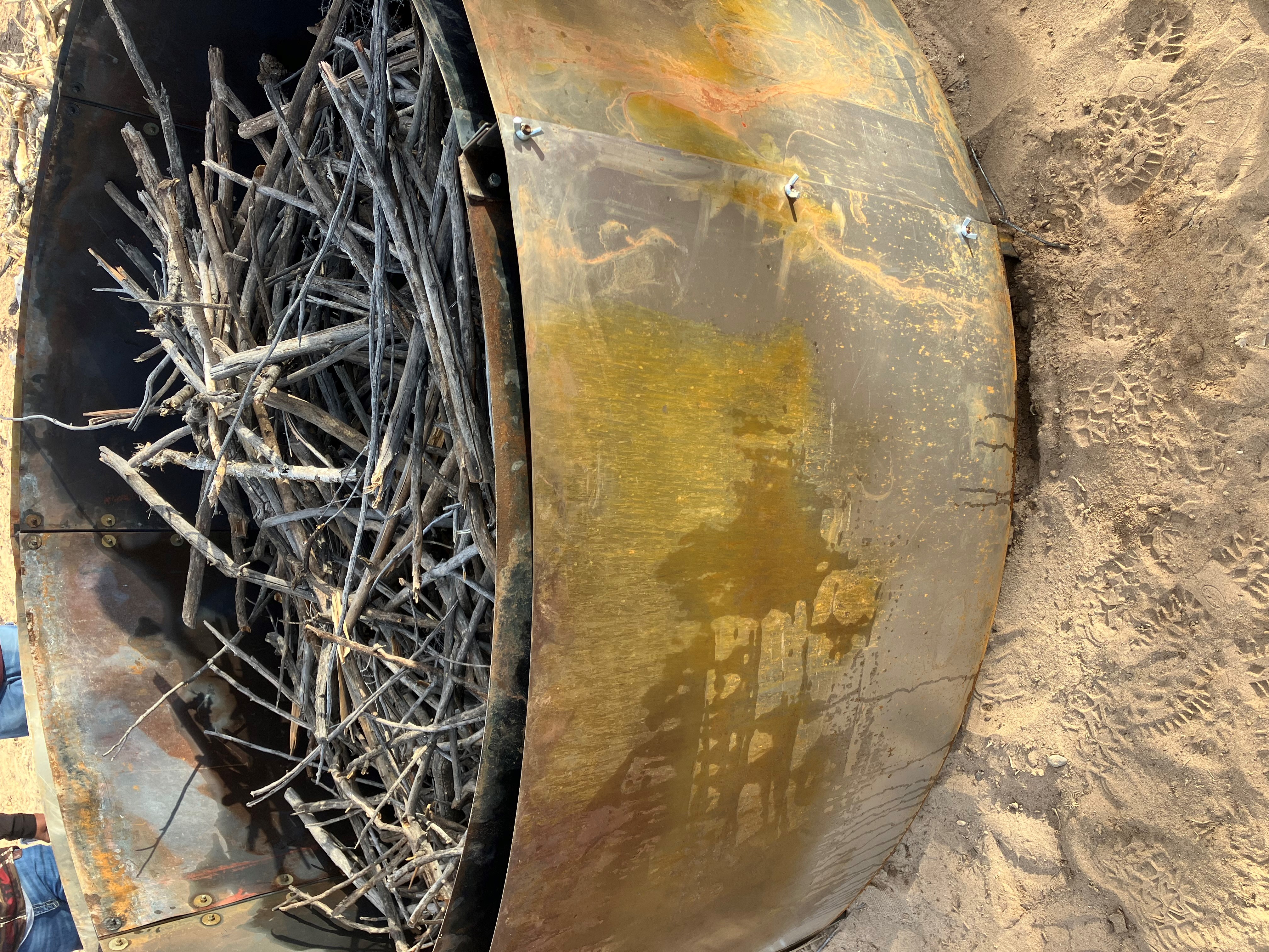Large metal fire pit filled with dry sticks and branches on sandy ground.