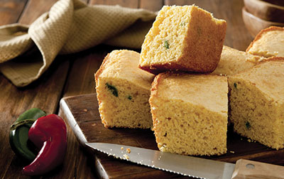 Photograph of pieces of cornbread on a cutting board with a knife and two chile peppers.