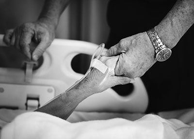 Photograph of a person in a hospital bed holding another person’s hand.