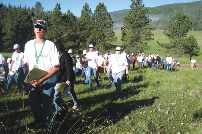 A group of 4-H WHEP participants hiking through a lush green field, dressed in jeans, white shirts, hats, and sunglasses. In the background, tall pine trees and distant mountains are visible. The participants are heading towards the contest site to evaluate the habitat for their team's wildlife management plan.