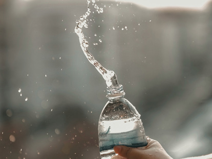 Hand holding a plastic water bottle as water splashes upward in the sunlight.