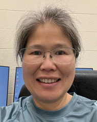 A smiling woman with gray hair and glasses takes a selfie in front of computer screens.