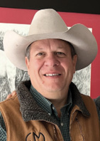 A smiling man wearing a cowboy hat and a brown vest, posing in front of a red backdrop.