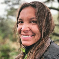 A smiling young woman with long, wavy hair, standing outdoors with a natural background.