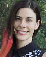 A smiling woman with long, dark hair featuring a gradient of reddish tones, wearing a black collared shirt with a patterned design.