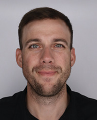 Close-up headshot of a smiling man with short brown hair and light stubble against a plain gray background.