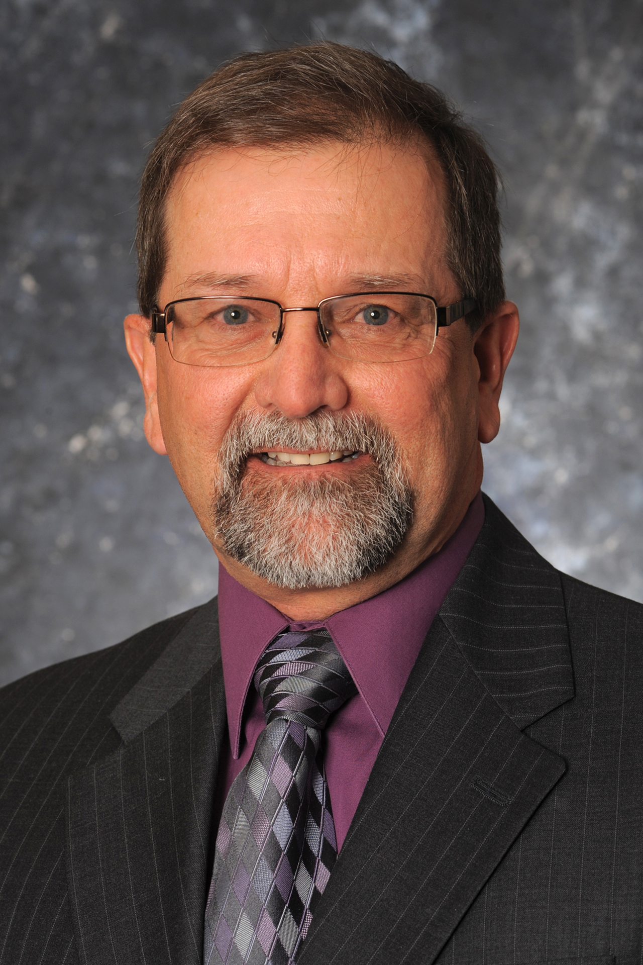 Professional headshot of a man with glasses, wearing a dark suit and tie, smiling against a blurred gray background.