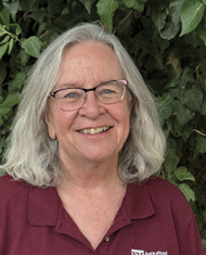 Smiling older woman with shoulder-length gray hair and glasses, wearing a maroon collared shirt, standing in front of leafy greenery.