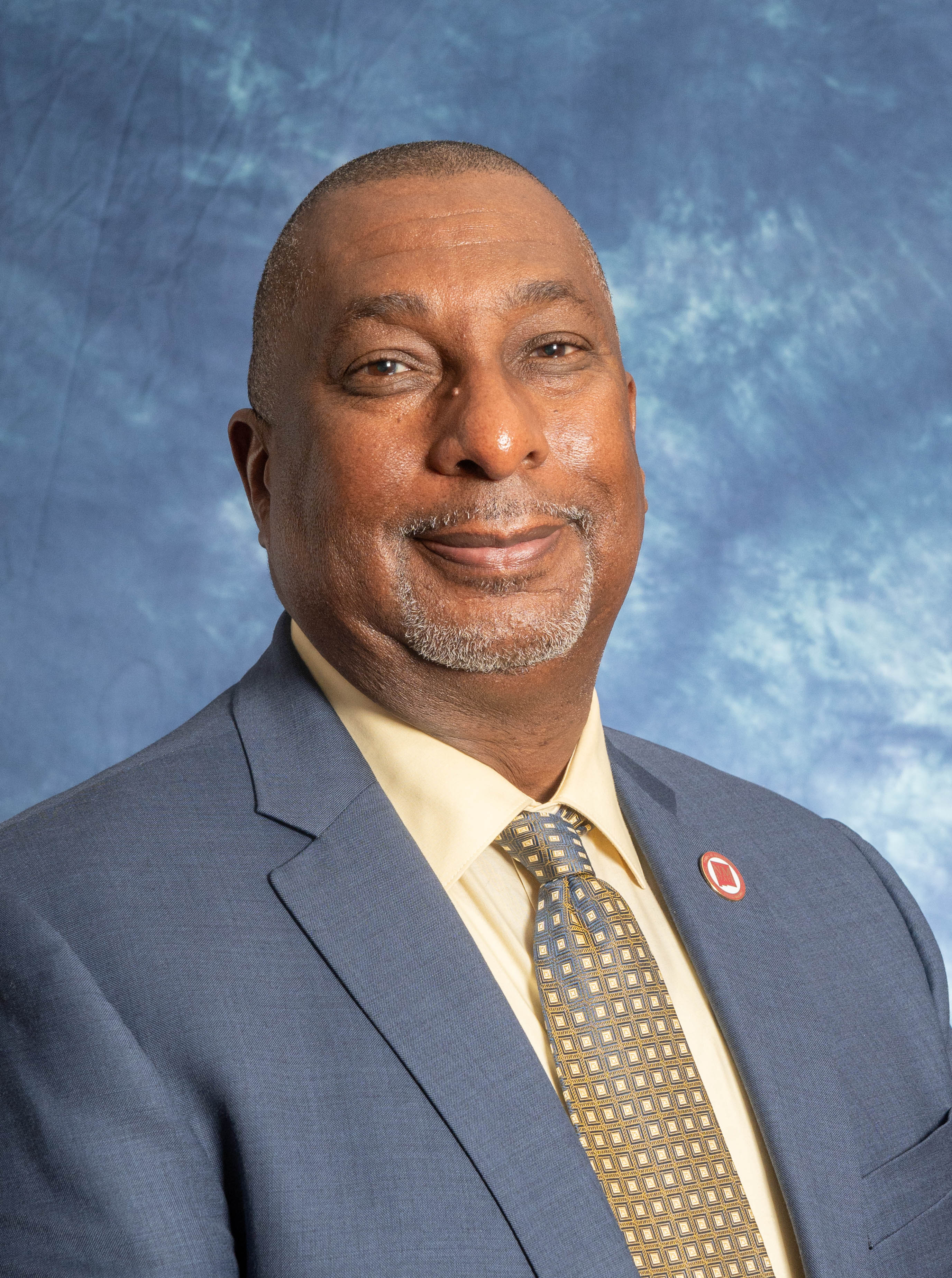 Professional headshot of a smiling man in a blue suit and patterned tie against a mottled blue studio backdrop.